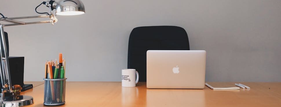 Modern home office workspace with MacBook laptop, coffee mug, pencil holder with colored pens, adjustable desk lamp, black office chair, and smartphone on wooden desk